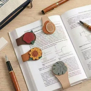 May include: Four leather magnetic bookmarks shaped like tabs, each featuring a distinct embossed design: a red rose, a yellow sunflower, a green monstera leaf, and a tan circular emblem. These bookmarks are placed on the pages of an open textbook.