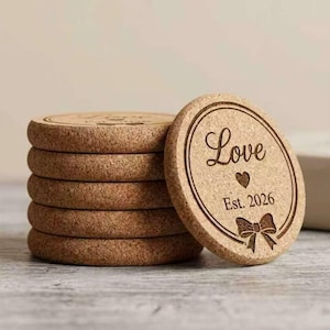 May include: A stack of six round cork coasters. One coaster is leaning against the stack, displaying the word "Love", a heart, the year "Est. 2026", and a bow design. The coasters are light brown.