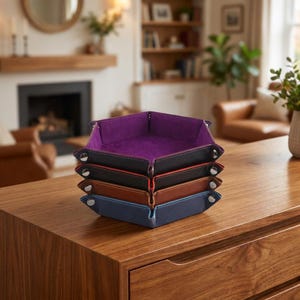 May include: A stack of five leather trays in various colors: purple, black, red, brown, and blue. Each tray has a contrasting interior and is secured with metal snaps. The trays are displayed on a wooden surface.