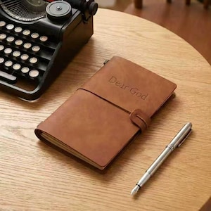 May include: A brown leather journal with the embossed text "Dear God" is secured with a strap. A silver fountain pen rests beside the journal on a wooden surface. A vintage typewriter is in the background.