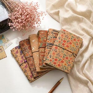 Puede incluir: Una colección de diarios con cubiertas de corcho con varios diseños, incluyendo mariposas, patrones geométricos, edificios e impresiones florales. Cada diario está encuadernado con un cierre elástico negro. Los diarios están dispuestos sobre una superficie blanca con un bolígrafo y flores secas.