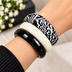 Puede incluir: Un conjunto de tres brazaletes gruesos usados juntos. El brazalete superior presenta un patrón ondulado en blanco y negro similar a una cebra. El brazalete central es de color blanco roto sólido. El brazalete inferior tiene un acabado negro brillante.