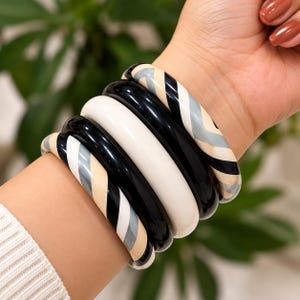 Può includere: Un set di cinque bracciali rigidi che combina modelli in nero tinta unita, bianco tinta unita e versioni a fantasia. I bracciali a fantasia presentano strisce diagonali che alternano i colori nero, bianco e grigio chiaro.