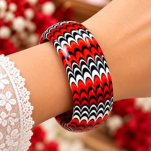 Puede incluir: Una pulsera rígida, gruesa y redondeada con un patrón repetitivo de ondas en rojo vibrante, negro intenso y blanco nítido. La superficie brillante muestra un diseño ondulado continuo que envuelve toda la circunferencia del accesorio.