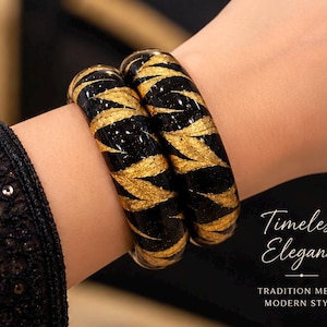 May include: Two black bangle bracelets featuring a gold leaf pattern with a glossy finish. The text Timeless Elegance TRADITION MEETS MODERN STYLE is visible in the lower right corner of the image. The bracelets have a smooth, rounded surface.