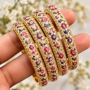 May include: Four gold-tone bangles featuring a repeating floral pattern with pink, purple, and blue enamel petals. The surface of each bangle is densely encrusted with small, clear, sparkling stones set against a metallic gold-tone base.