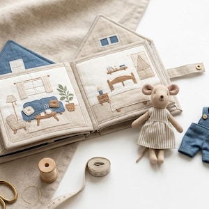 May include: A fabric house-shaped book with embroidered interior scenes, including a living room and bedroom. A small stuffed mouse doll stands next to the book, accompanied by a pair of blue overalls and a floral dress. Gold scissors and a spool are nearby.