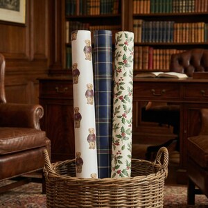 Pu&ograve; includere: Tre rotoli di carta da regalo decorativa in un cesto di vimini. I rotoli presentano motivi di un orsacchiotto, quadri blu navy e bacche di agrifoglio. Lo sfondo include un ambiente di biblioteca con poltrone in pelle e una scrivania in legno.