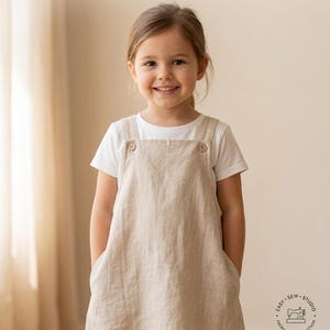 May include: A young girl wearing a beige pinafore dress over a white t-shirt. The image features text that reads "Girls Pinafore Dress Pattern" and indicates the pattern is for 14 sizes, from 12 months to 16 years, and is beginner-friendly. Digital download available.