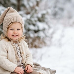 May include: A child wearing a beige knit hat with a long pointed tip and a matching cardigan. The child is smiling while sitting on a wooden bench in a snowy, wintery scene. The hat has ear flaps and ties.