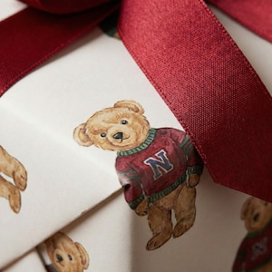 Pu&ograve; includere: Una confezione regalo avvolta in carta bianca con un motivo ripetuto di orsetti di peluche con maglioni color amaranto con finiture verdi e bianche e la lettera "N". La scatola &egrave; legata con un ampio nastro di raso color amaranto.