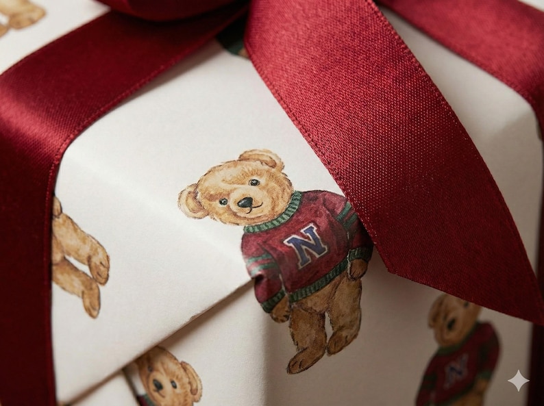 Pu&ograve; includere: Una confezione regalo avvolta in carta bianca con un motivo ripetuto di orsetti di peluche con maglioni color amaranto con finiture verdi e bianche e la lettera "N". La scatola &egrave; legata con un ampio nastro di raso color amaranto.