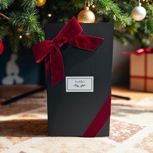 May include: A black gift box with a red velvet ribbon and bow, placed under a decorated Christmas tree. The box has a small white label with the word "SoHo" and a sprig design. The box is on a patterned rug.