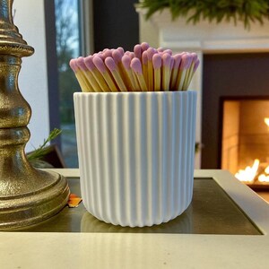 May include: A white, ribbed ceramic match holder filled with matches. The matches have pink tips and are arranged in a neat bundle. The holder is on a table with a gold candlestick and a fireplace in the background.