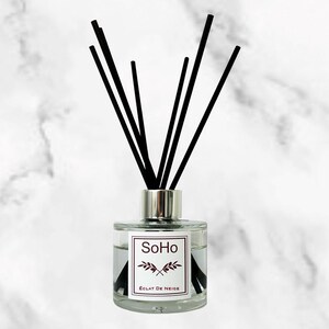 May include: A clear glass diffuser bottle with black reeds. The bottle has a silver cap and a white label with the text "SoHo" and "ÉCLAT DE NEIGE". The reeds are arranged in a fan shape, creating a decorative and aromatic home fragrance.