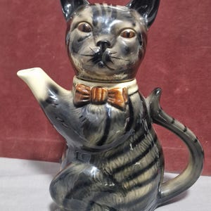 May include: A ceramic cat-shaped teapot with a grey and black striped design. The cat has a brown bow tie and a white collar. The teapot has a spout and a handle. The cat is sitting upright.