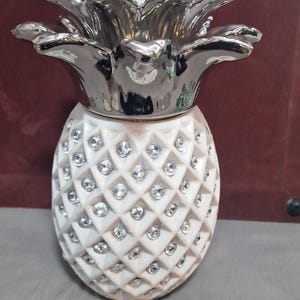 May include: A decorative pineapple ornament. The white body is studded with crystals, and the top is silver-coloured. This ornament is likely for home decor. The item is designed to resemble a pineapple.