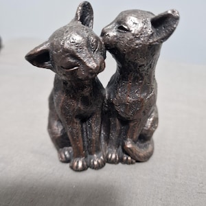 May include: A bronze-effect sculpture of two cats, one nuzzling the other. The cats are depicted with closed eyes, suggesting affection. The sculpture has a textured finish and is a decorative piece.