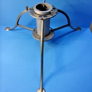 May include: A metallic silver Christmas tree stand featuring a central cylindrical tube for holding a tree trunk, supported by three curved legs with flat circular feet and three adjustable metal screws at the top to secure the tree in place.