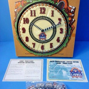 May include: A Lionel 100th Anniversary limited edition train clock featuring a cream-colored face with red numbers, a black train track border, and a mountain scene top. Includes operating instructions, a certificate, and a small train set.