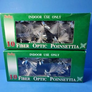 May include: Two stacked green rectangular boxes labeled 10 Fiber Optic Poinsettia. Each box features a clear window displaying white fiber optic floral decorations with silver accents. The text reads Sterling, Indoor Use Only, and 10 Fiber Optic Poinsettia.