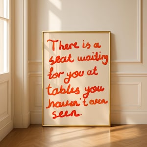 May include: A framed art print with the text "There is a seat waiting for you at tables you haven't even seen" in orange script on a cream background. The frame is gold, and the print is displayed on a wooden floor.