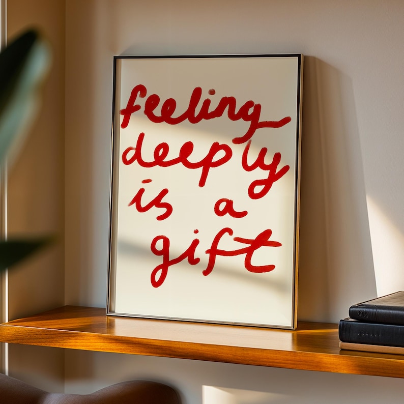 May include: A framed art print with the words "feeling deeply is a gift" in red script on an off-white background. The frame is gold-colored, and the print is displayed on a wooden shelf.