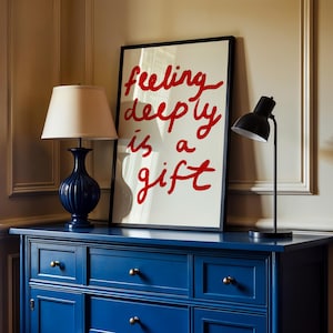 May include: A navy blue dresser with brass knobs supports a framed art print and two lamps. The art print reads "feeling deeply is a gift" in red script. A blue lamp with a beige shade and a black desk lamp are on the dresser.