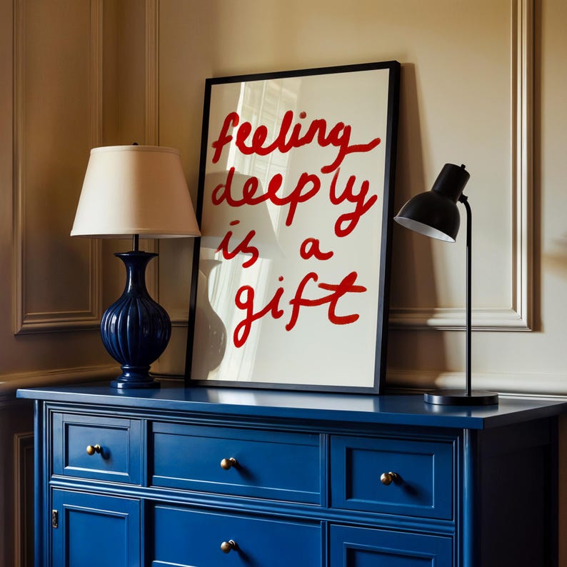 May include: A navy blue dresser with brass knobs supports a framed art print and two lamps. The art print reads "feeling deeply is a gift" in red script. A blue lamp with a beige shade and a black desk lamp are on the dresser.
