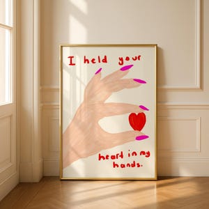 May include: A framed art print featuring a hand holding a red heart. The hand has pink fingernails and is painted in a light skin tone. The text "I held your heart in my hands" is written in red.