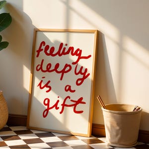 May include: A framed art print with the words "feeling deeply is a gift" in red script. The artwork has a cream-colored background and a natural wood frame. The print leans against a wall next to a beige bucket.