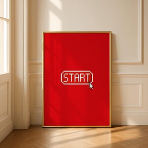 Könnte beinhalten: Ein gerahmter Kunstdruck mit leuchtend rotem Hintergrund. Der Druck zeigt das Wort "START" in einer pixeligen weißen Schrift, eingeschlossen in einem weißen Rechteck. Ein weißer Cursor zeigt auf das Wort. Der Rahmen ist goldfarben.