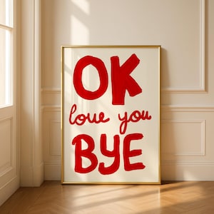 Puede incluir: Un cuadro enmarcado con las palabras "OK love you BYE" en letras rojas sobre un fondo blanquecino. El marco dorado añade un toque de elegancia a la obra de arte, que está apoyada contra una pared.