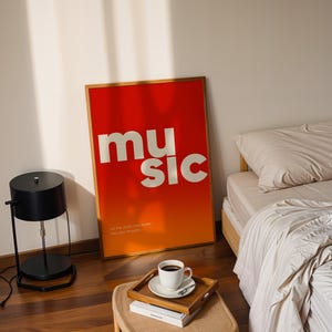 Puede incluir: Un p&oacute;ster enmarcado con la palabra "music" en letras blancas sobre un fondo degradado rojo y naranja. El p&oacute;ster est&aacute; apoyado contra una pared cerca de una cama y una l&aacute;mpara negra. Una bandeja con una taza de caf&eacute; est&aacute; cerca.