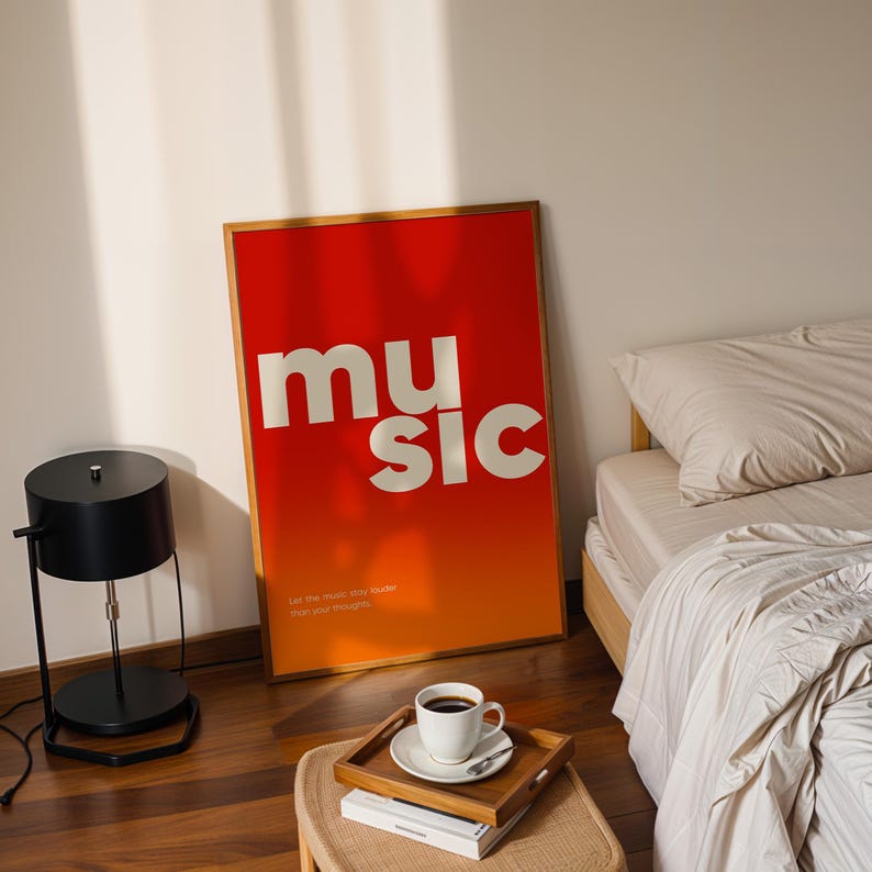 Puede incluir: Un p&oacute;ster enmarcado con la palabra "music" en letras blancas sobre un fondo degradado rojo y naranja. El p&oacute;ster est&aacute; apoyado contra una pared cerca de una cama y una l&aacute;mpara negra. Una bandeja con una taza de caf&eacute; est&aacute; cerca.