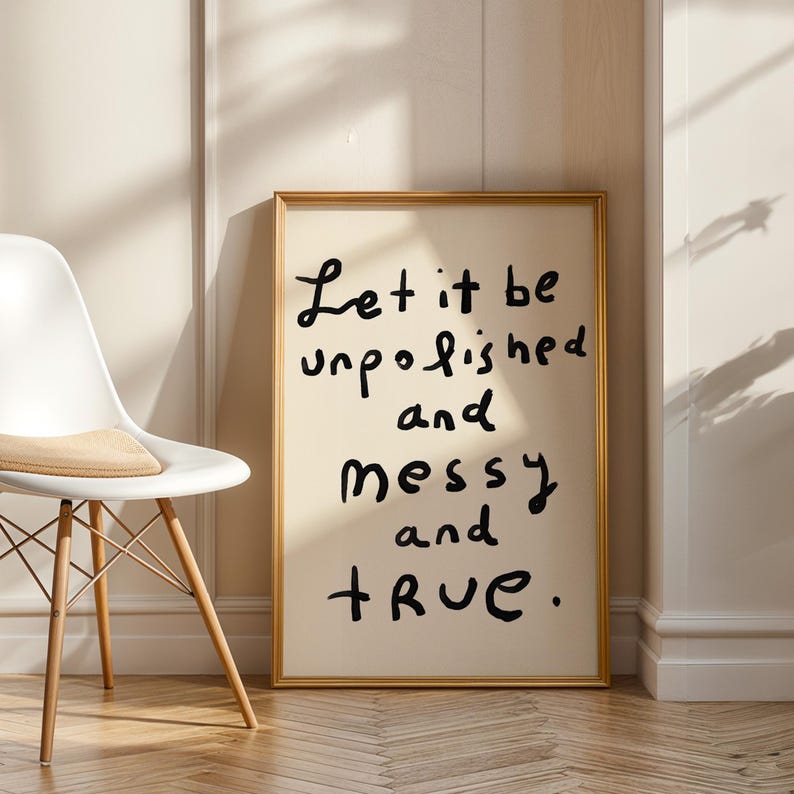 May include: A framed art print with the words "Let it be unpolished and messy and TRUE" in black handwritten font. The print has a cream-colored background and a gold frame. A white chair with wooden legs is to the left.