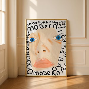 Puede incluir: Una impresión artística enmarcada que presenta un rostro estilizado con ojos azules y piel color melocotón. La obra de arte incluye la palabra "MODERN" y otro texto en negro. El marco es dorado.