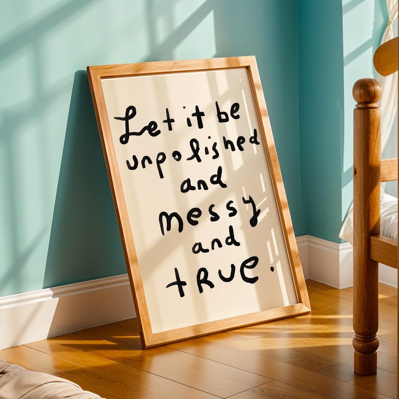 May include: A framed art print with a light beige background and a wooden frame. The black handwritten text reads "Let it be unpolished and messy and true!" The artwork leans against a light blue wall.