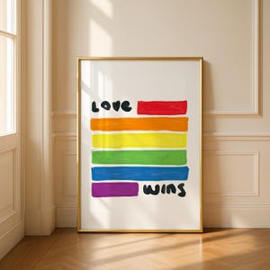 Puede incluir: Una impresión artística enmarcada con un fondo blanco y un diseño de arcoíris. Las palabras "Love" y "Wins" están escritas en negro. La obra de arte se muestra en un marco dorado y se apoya contra una pared.