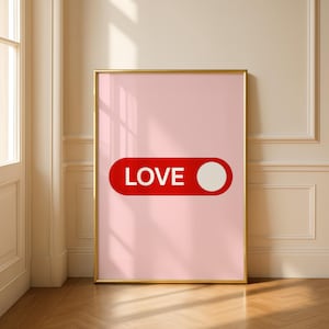以下が含まれることがあります： ピンク色の背景に、赤い長方形が描かれたフレーム入りアートプリント。 白い文字で「LOVE」と書かれ、長方形の右側には白い円があります。 フレームは金色です。