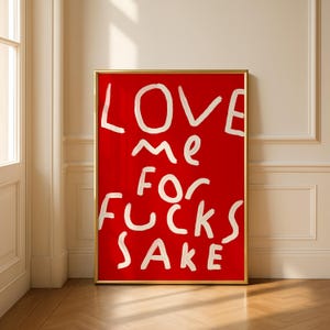May include: A framed art print with a bold red background. The white text reads "LOVE me FOR FUCKS SAKE" in a handwritten style. The frame is gold, and the artwork is displayed against a neutral wall and wooden floor.