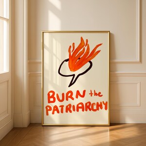 May include: A framed art print with a gold border. The artwork features a black speech bubble with orange flames and the words "BURN the PATRIARCHY" in orange, handwritten-style lettering. The background is a cream color.