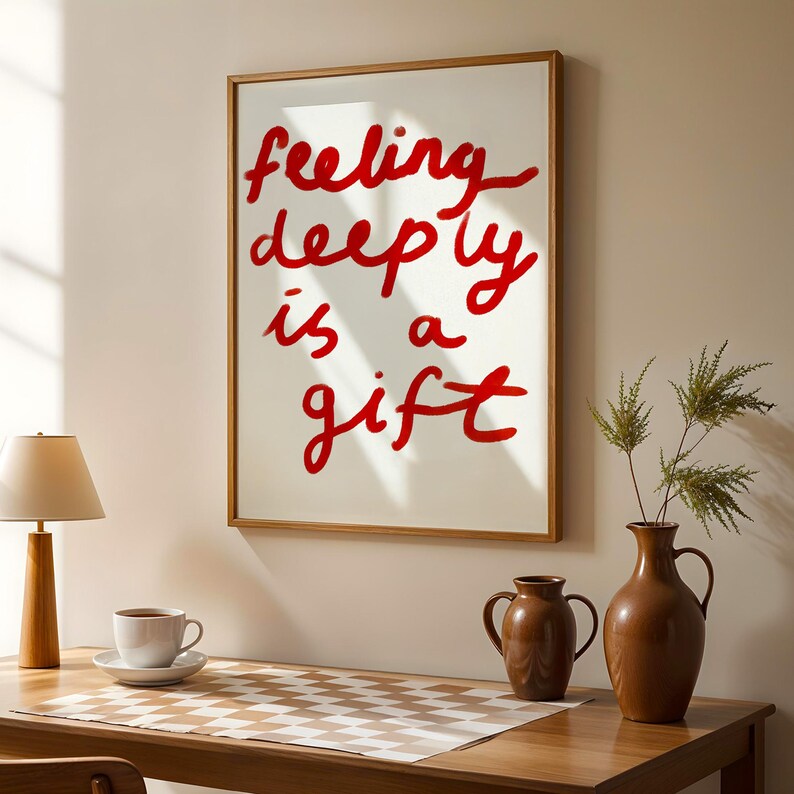 May include: A framed art print with the words "feeling deeply is a gift" in red script on a white background. The frame is a light brown color. The print is displayed on a wooden table with a lamp, coffee cup, and vases.