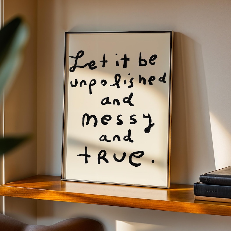 May include: A framed print with the handwritten text "Let it be unpolished and messy and true." The black text is on a cream-colored background, framed in a thin gold frame. The print rests on a wooden shelf.
