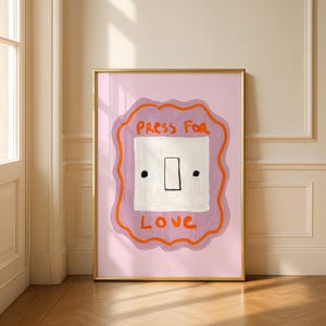 Op de afbeelding: Een ingelijste kunstprint met een illustratie van een lichtschakelaar. Het kunstwerk heeft een roze achtergrond, een oranje omtrek en een paarse vierkant. De witte schakelaar heeft de woorden "Press for Love" in oranje. De lijst is goud.