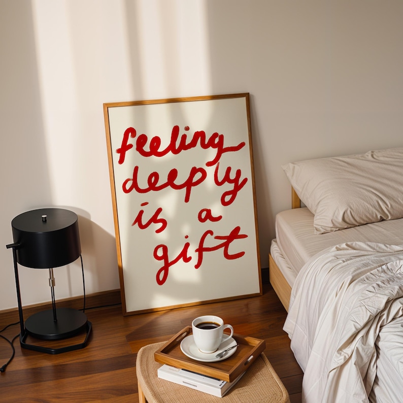 May include: A framed art print with the words "feeling deeply is a gift" in red script. A black table lamp, a wooden tray with a coffee cup, and a bed with beige bedding are also visible.