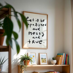 May include: A framed print with the words "Let it be unpolished and messy and true" in black handwritten font. The print is in a light wood frame. Two smaller framed pictures and a potted plant sit below.
