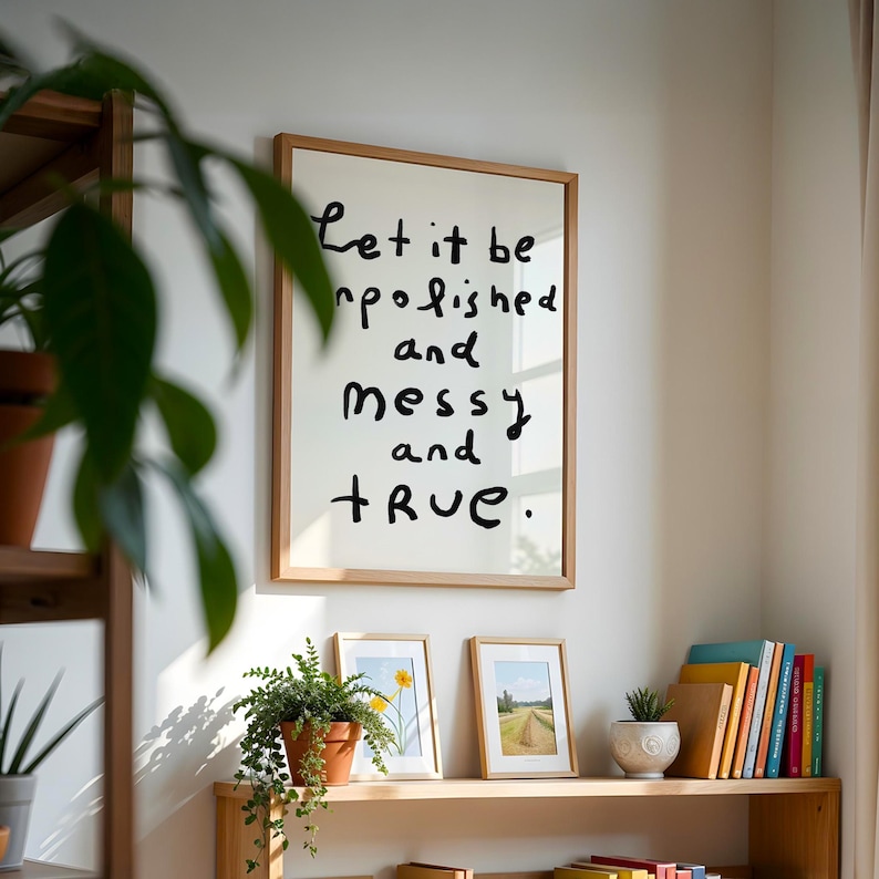May include: A framed print with the words "Let it be unpolished and messy and true" in black handwritten font. The print is in a light wood frame. Two smaller framed pictures and a potted plant sit below.