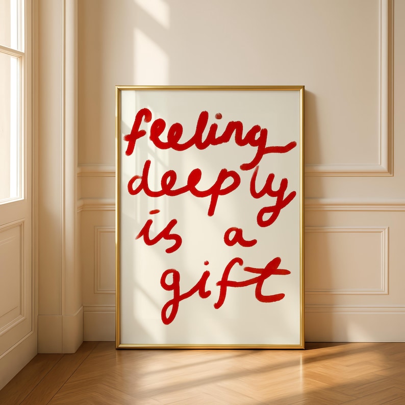 May include: A framed art print with the words "feeling deeply is a gift" in red cursive script on an off-white background. The frame is gold, and the artwork is displayed against a neutral wall and wooden floor.