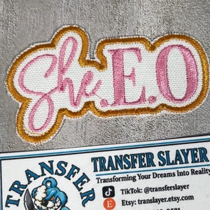 May include: An embroidered patch featuring the text "She E.O" in pink script and block lettering with a white background and a gold border. Below the patch is a white business card with the text "TRANSFER SLAYER" and a blue cartoon bear logo.
